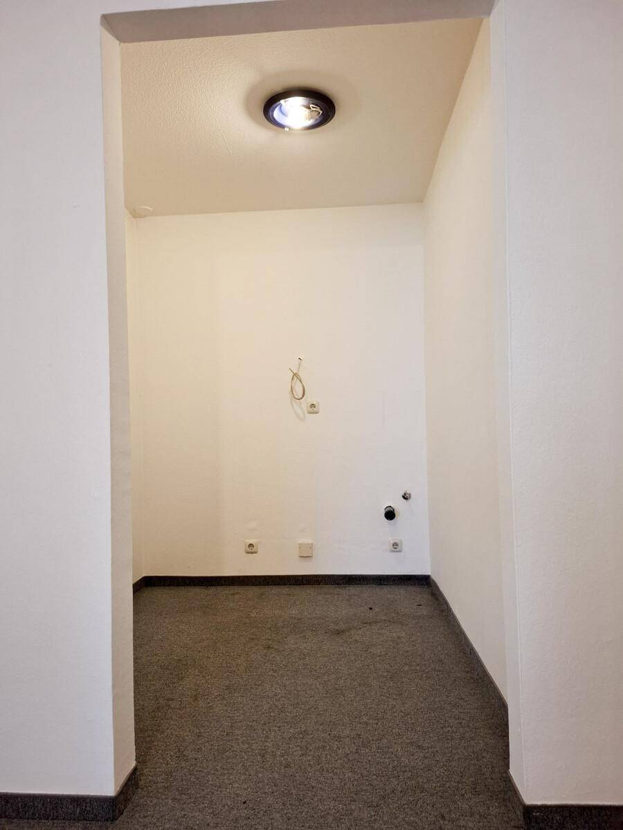Kleiner, leerer Raum mit grauem Teppichboden und sichtbaren Elektroinstallationen an der Wand.