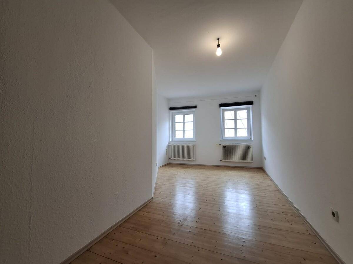 Leeres Zimmer mit zwei Fenstern, Holzboden und Heizkörpern unter den Fenstern.