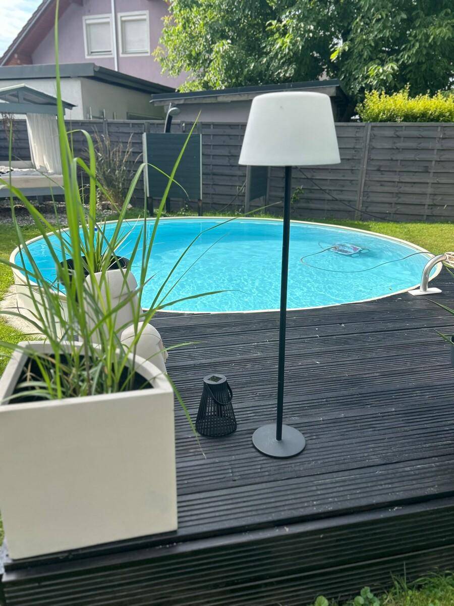 Gemütlicher Poolbereich im Garten mit Holzdeck, Stehlampe und Pflanzen für abendliche Beleuchtung.