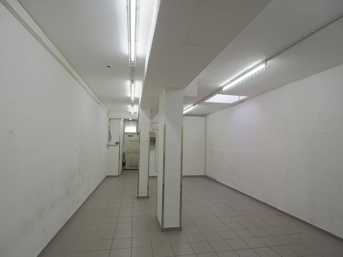Langer, leerer Gewerberaum mit Fliesenboden, weißen Wänden, Säulen und durchgehender Leuchtstoffbeleuchtung.