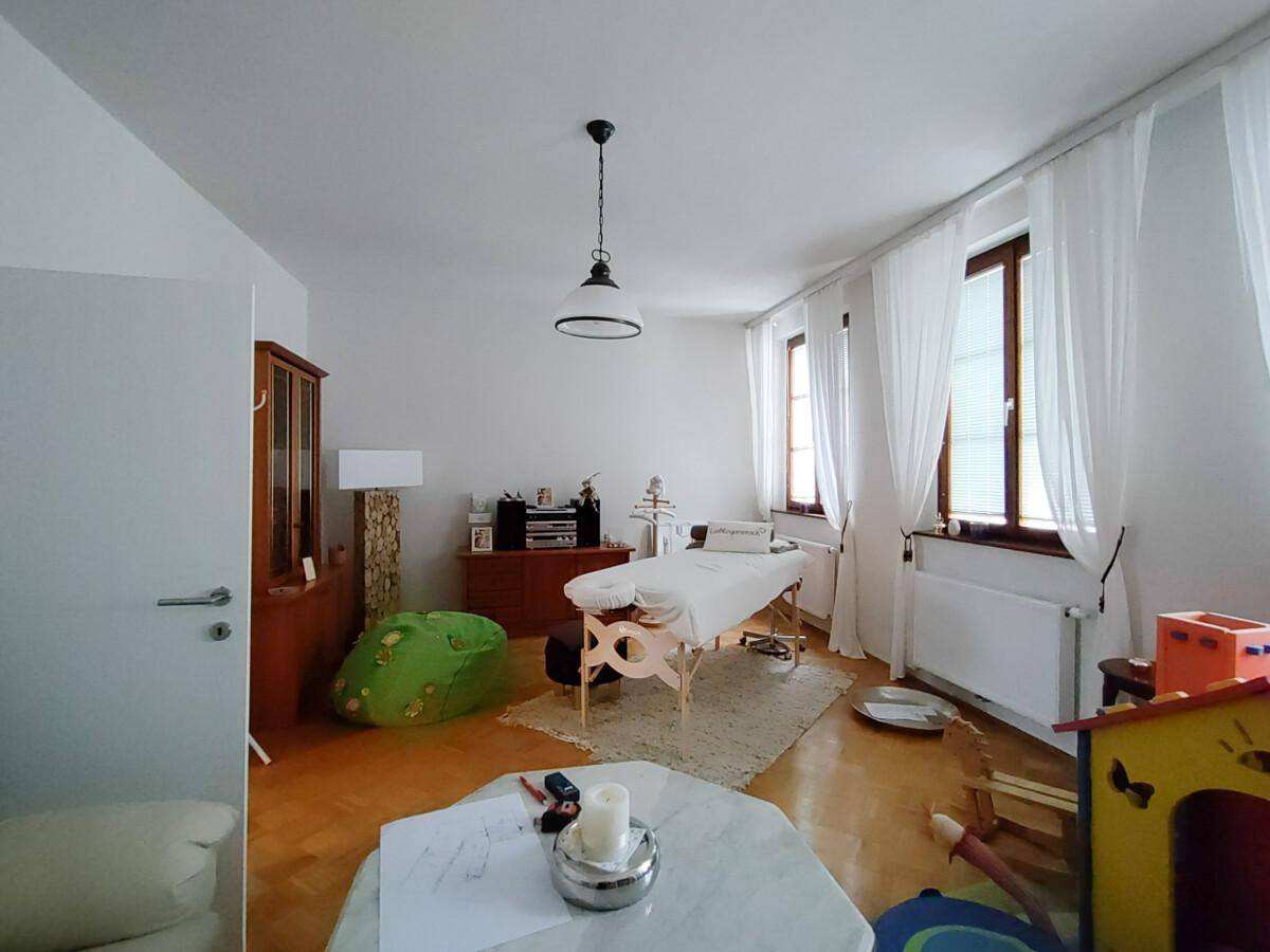 Heller Raum mit Massageliege, Holzmöbeln und großen Fenstern, vielseitig nutzbar.