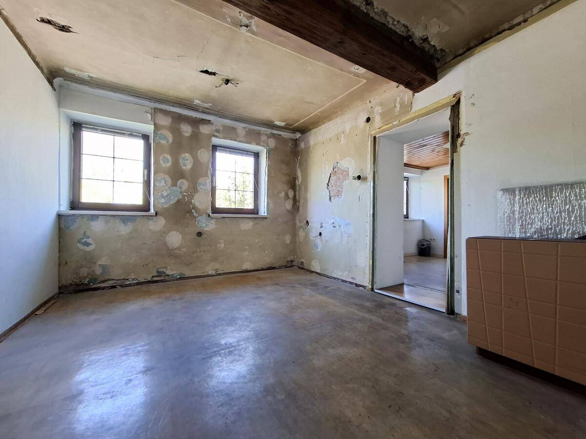 Renovierungsbedürftiger Raum mit freigelegten Wänden, zwei Fenstern und Holzbalken.