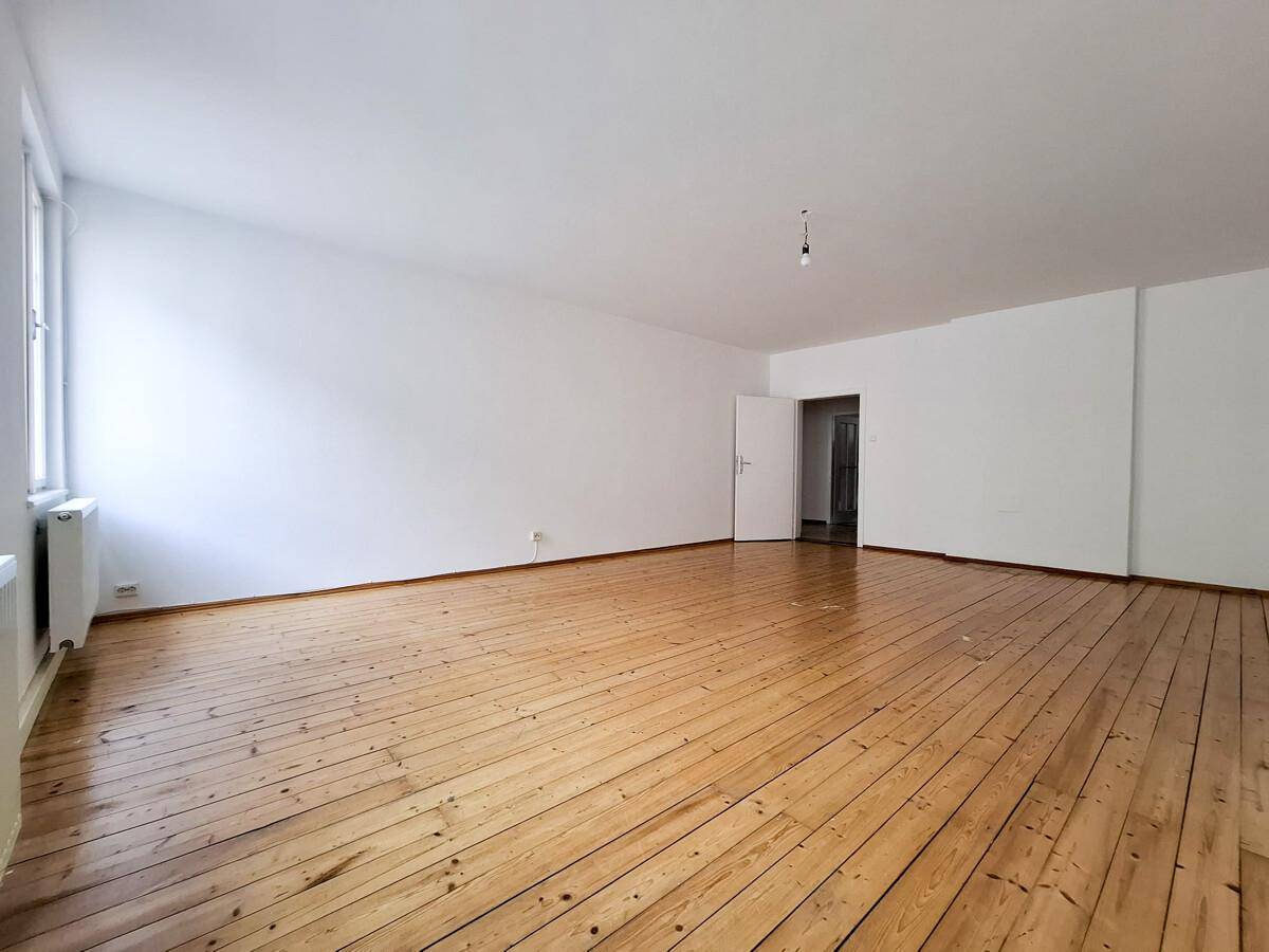 Geräumiges Zimmer mit Holzdielenboden, weißen Wänden und offenem Durchgang zu einem weiteren Bereich.