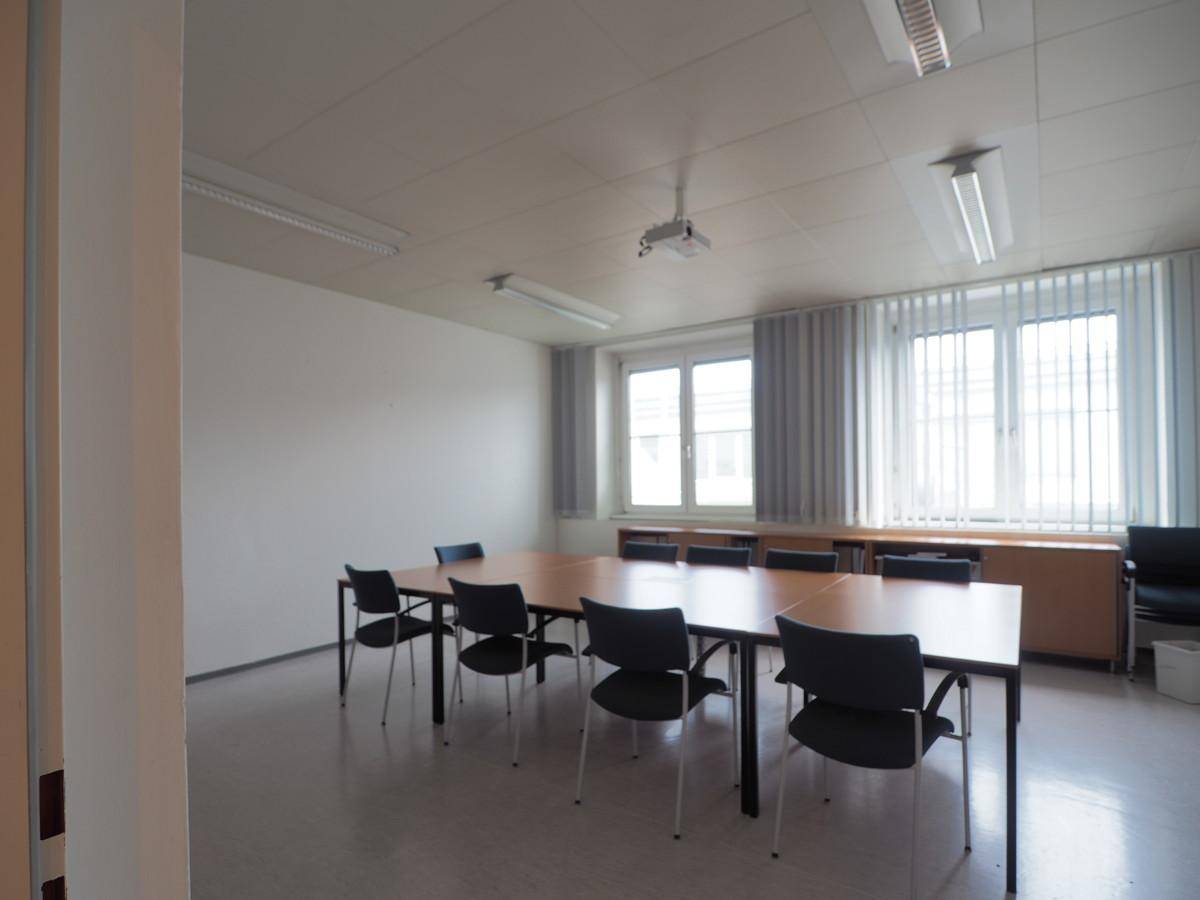 Funktionaler Konferenzraum mit mehreren Tischen, Stühlen und einem Projektor an der Decke.
