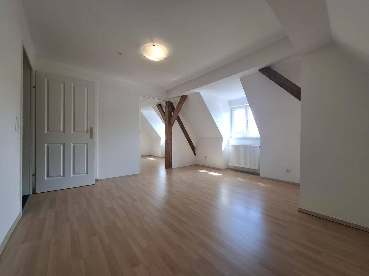 Helles Zimmer im Dachgeschoss mit Holzbalken, Parkettboden und schrägen Wänden.