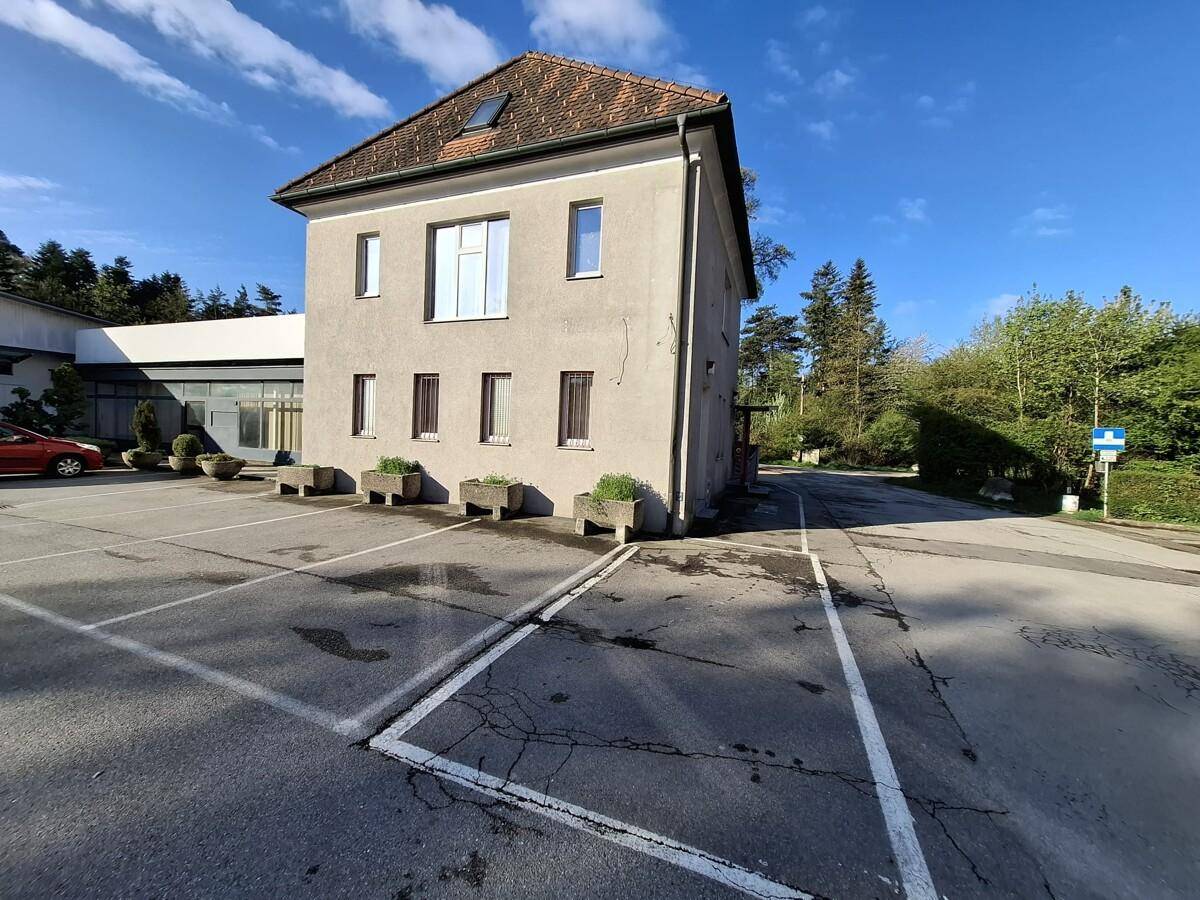 Gebäudeansicht mit Parkplätzen und Bäumen im Hintergrund, beleuchtet von der Morgensonne.