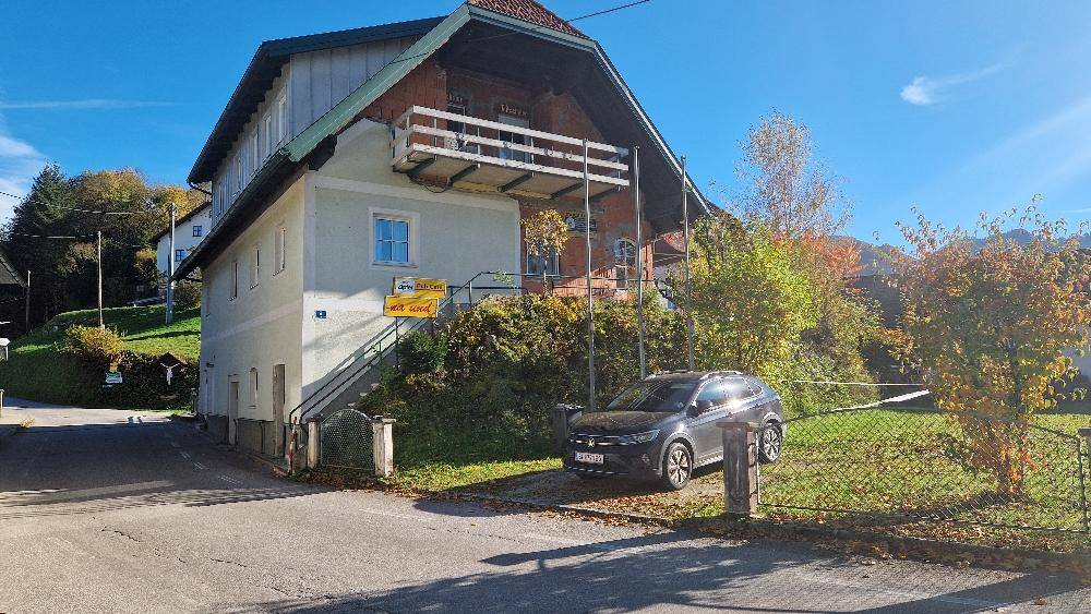Gebäudeansicht mit Balkon, Zufahrt und einem geparkten Auto vor grüner Umgebung.
