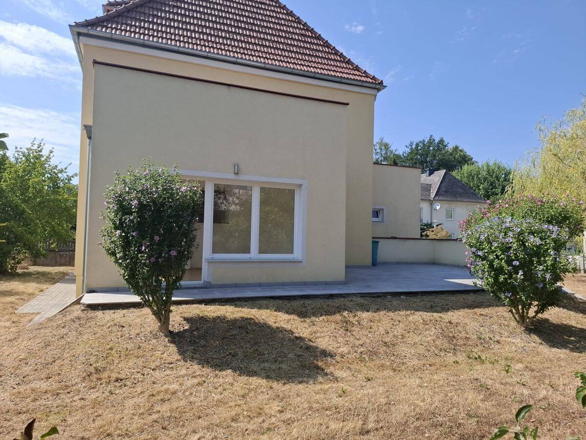 Seitenansicht des Hauses mit gepflastertem Bereich und trockenem Rasen unter blauem Himmel.