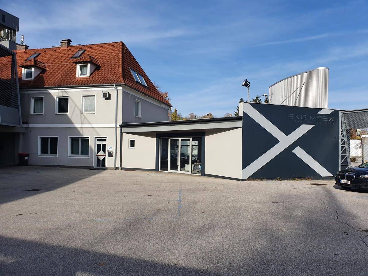 Rückansicht eines Geschäftsgebäudes mit einem großen Parkplatz und einem angrenzenden älteren Wohnhaus.