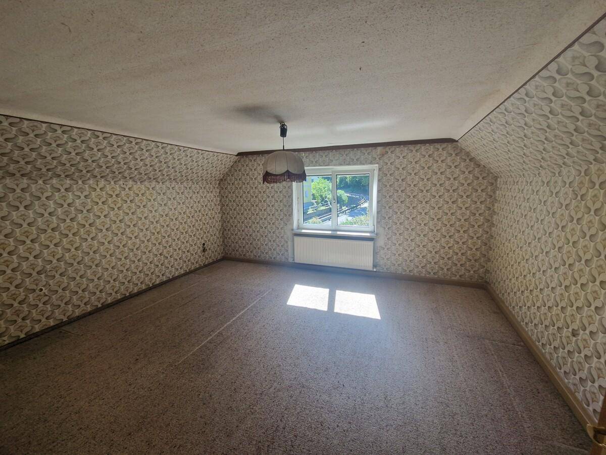 Dachzimmer mit gemusterter Tapete, Teppichboden und einem Fenster mit Blick ins Grüne.