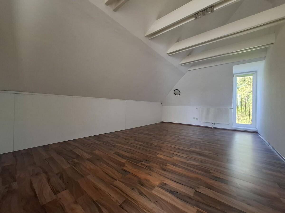 Geräumiger Dachgeschossraum mit Holzboden, Dachschräge und einem Fenster mit Blick ins Grüne.