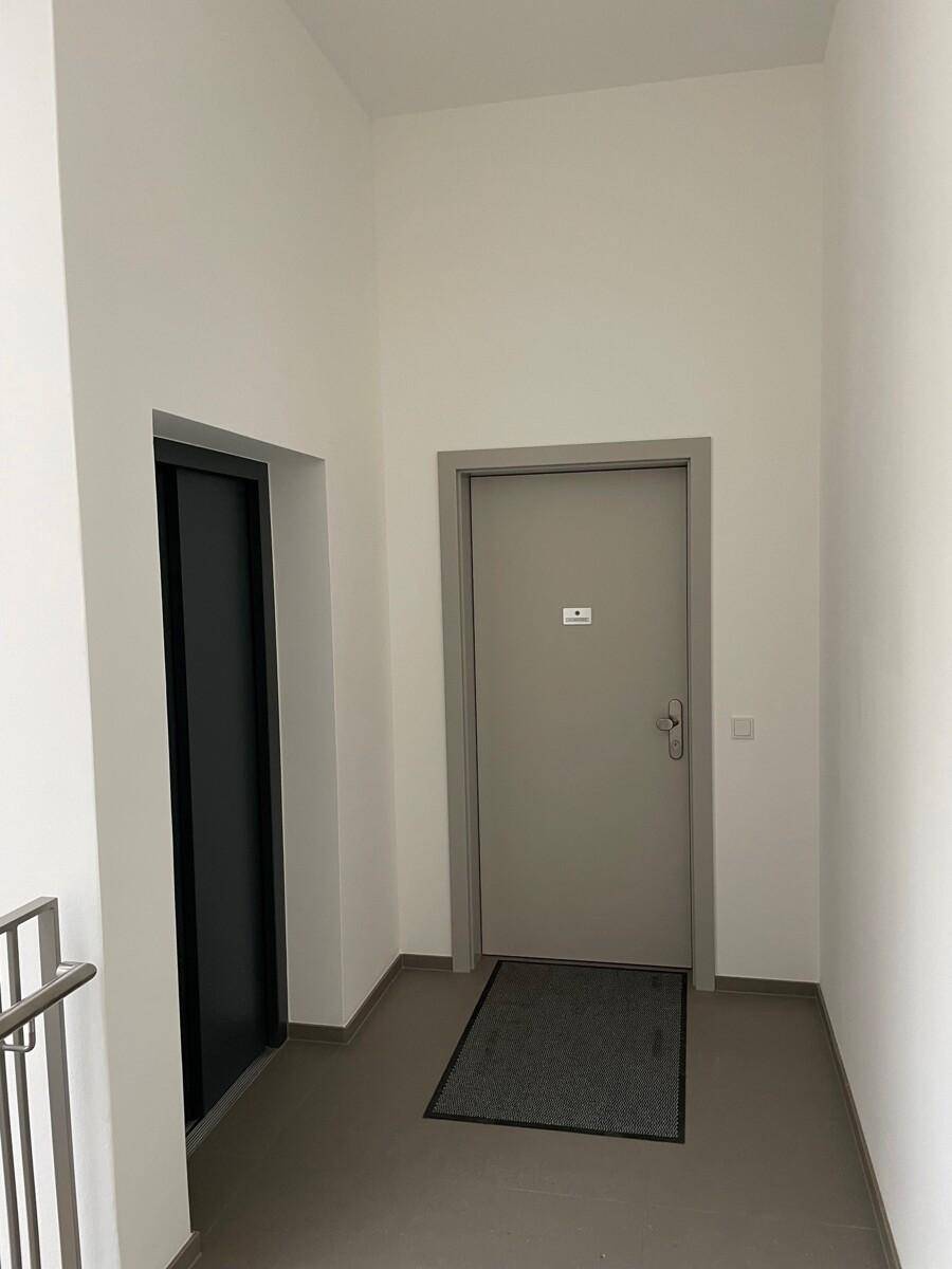 Heller Flur mit grauer Eingangstür und Zugang zu einem Aufzug.