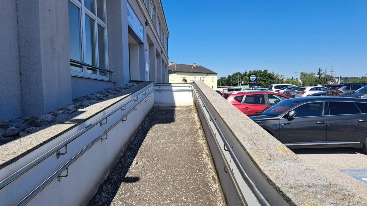 Barrierefreie Rampe mit Handläufen führt zum Eingang eines Gebäudes mit Parkplätzen.