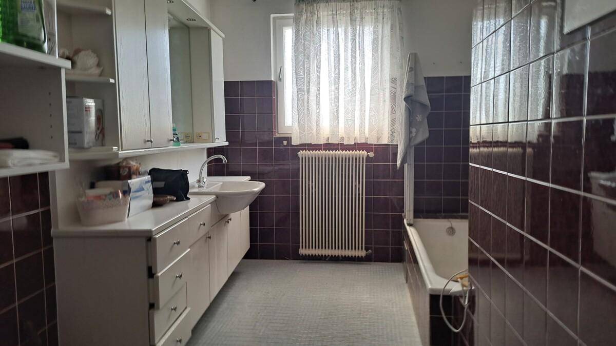 Badezimmer mit dunklen Fliesen, Badewanne, Waschbecken und Fenster, im Stil der 80er Jahre.