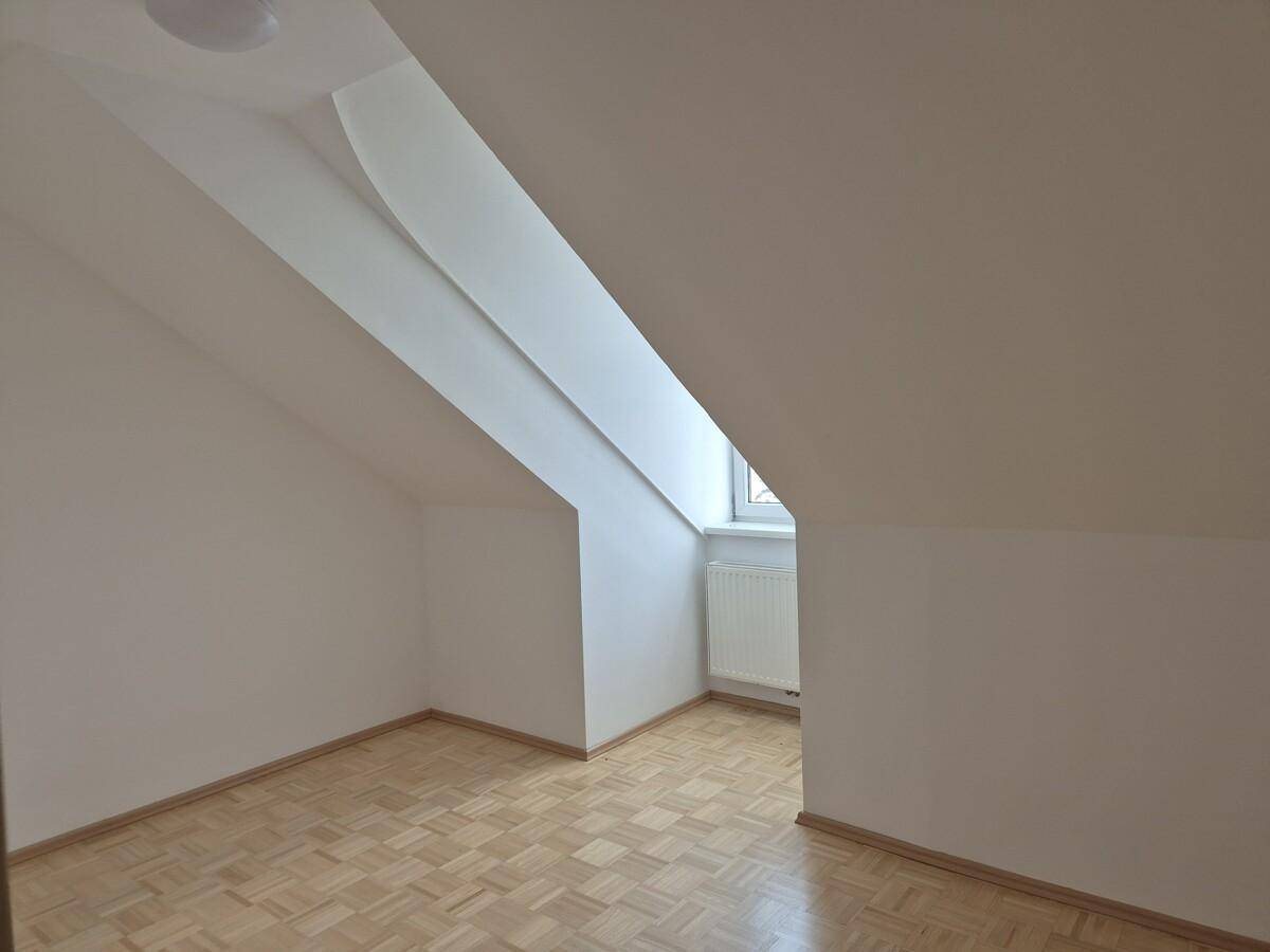 Dachzimmer mit schrägen Wänden, hellem Parkettboden und einem Fenster, das Tageslicht hereinlässt.
