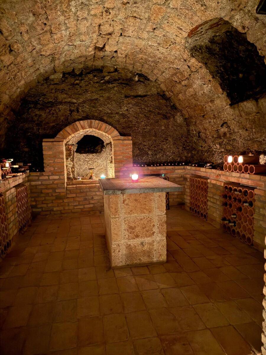 Historischer Weinkeller mit Ziegelgewölbe, Flaschenregalen und zentralem Steintisch, warm beleuchtet.