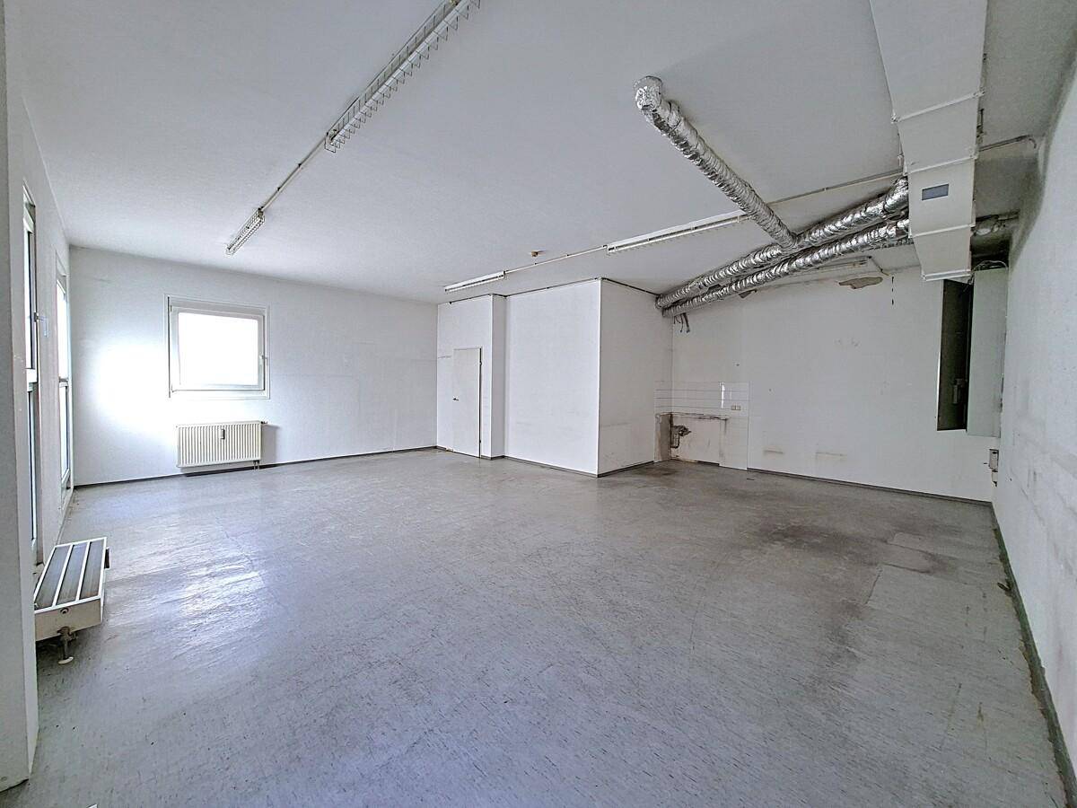 Lagerraum mit Betonboden, sichtbaren Lüftungsrohren und einem Fenster, funktional und geräumig.