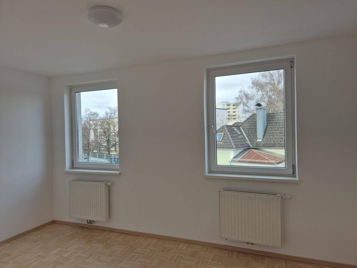 Heller Raum mit zwei Fenstern, Parkettboden und Blick auf die umliegenden Dächer und Bäume.