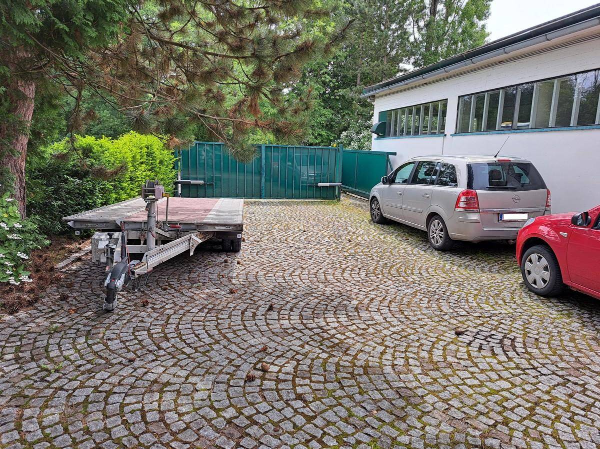 Gepflasterter Außenbereich mit Parkplätzen und einem Anhänger, umgeben von Bäumen und Grünflächen.