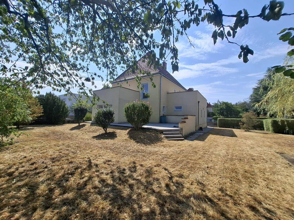 Rückansicht des Hauses mit trockenem Rasen und üppigem Grün im Hintergrund unter blauem Himmel.