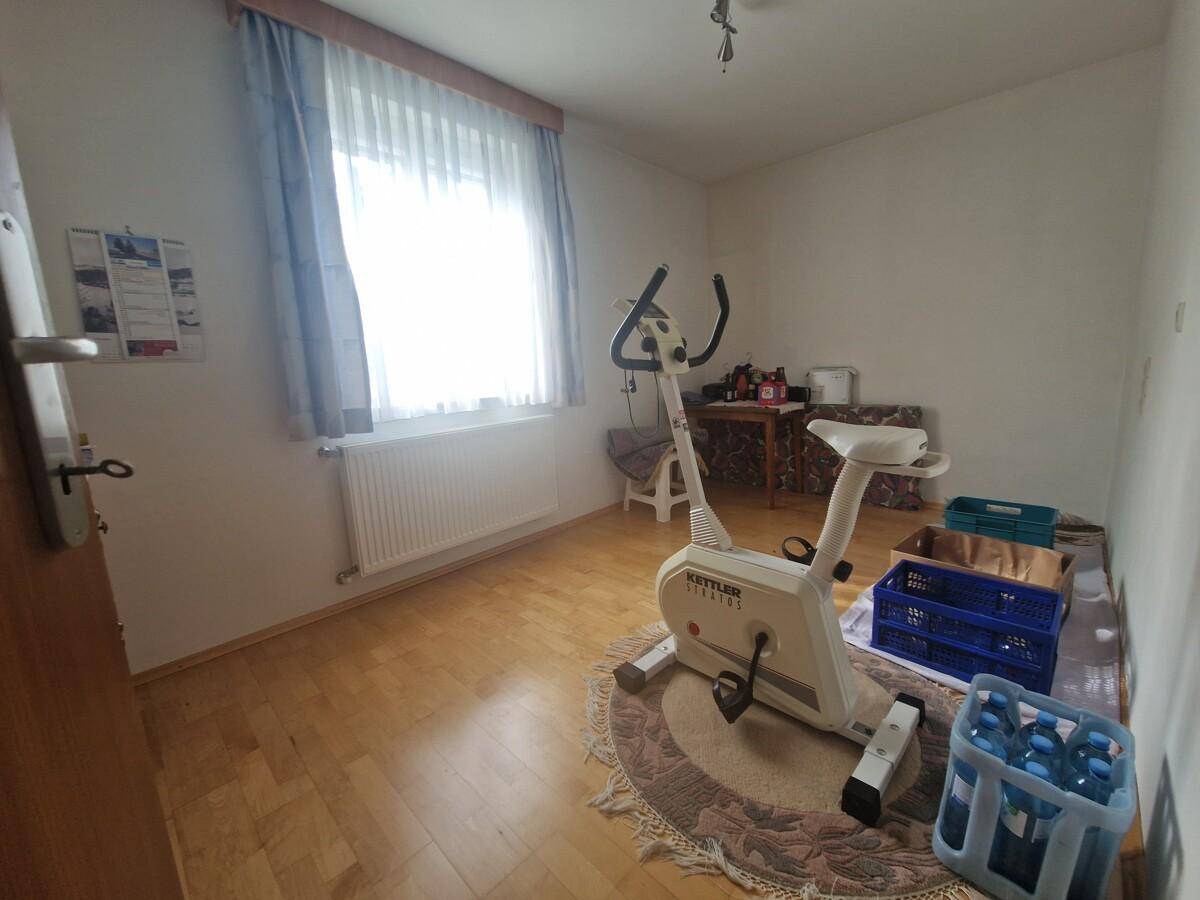 Helles Zimmer mit Parkettboden, Fenster und einem Heimtrainer, vielseitig nutzbar.