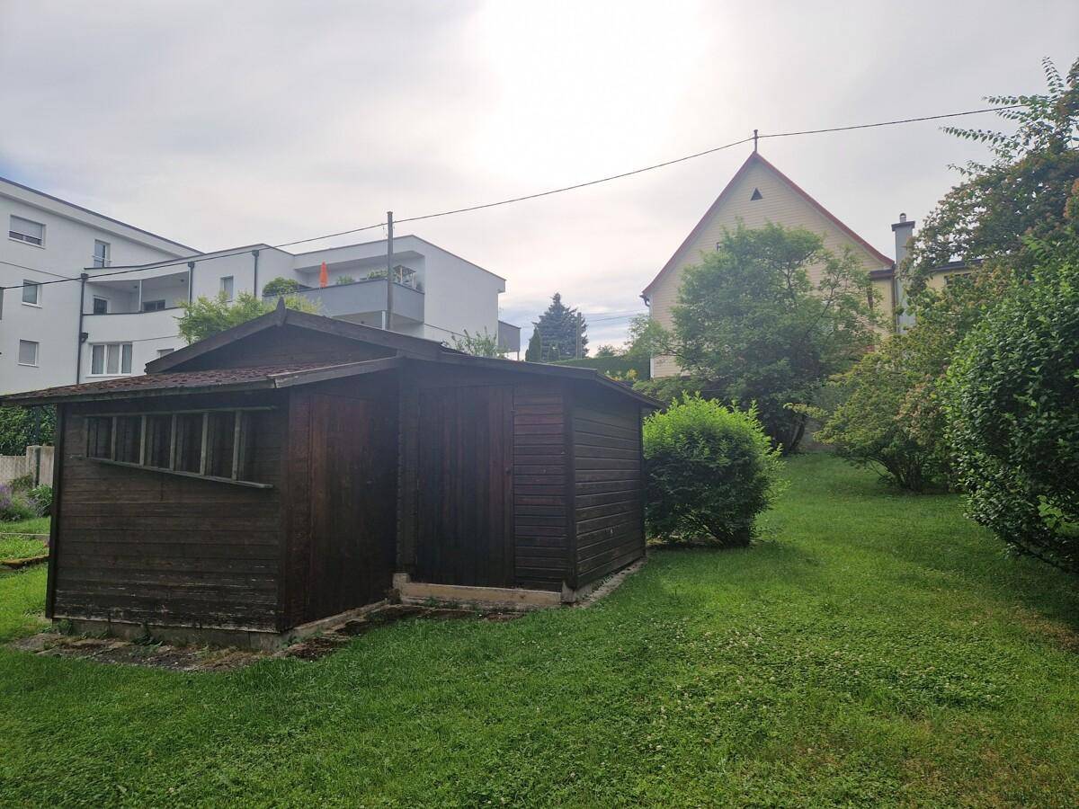 Kleiner Holzschuppen im grünen Garten, umgeben von Bäumen und Sträuchern.