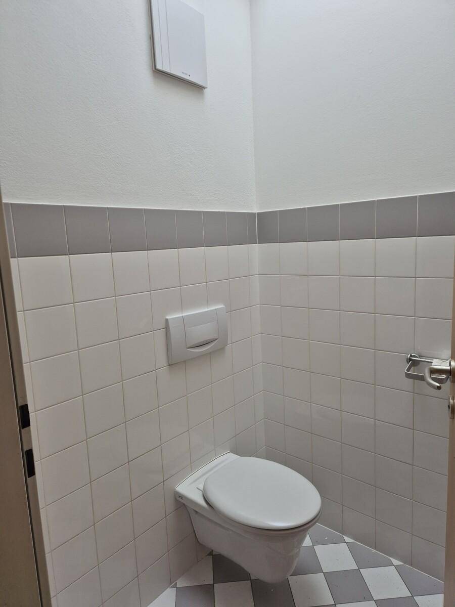 Modernes Gäste-WC mit wandhängender Toilette, weißen und grauen Fliesen sowie einem Abluftventilator.