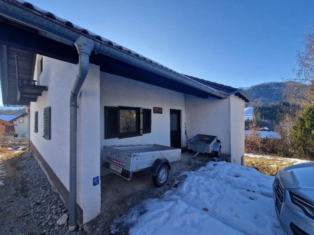 Hausfassade mit weißem Putz und überdachtem Bereich, davor stehen zwei Anhänger im Schnee.
