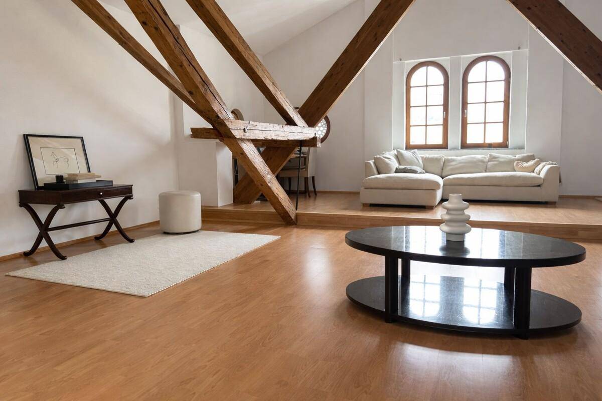 Helles Wohnzimmer im Dachgeschoss mit sichtbaren Holzbalken, Parkettboden und gemütlicher Sitzecke unter Rundbogenfenstern.