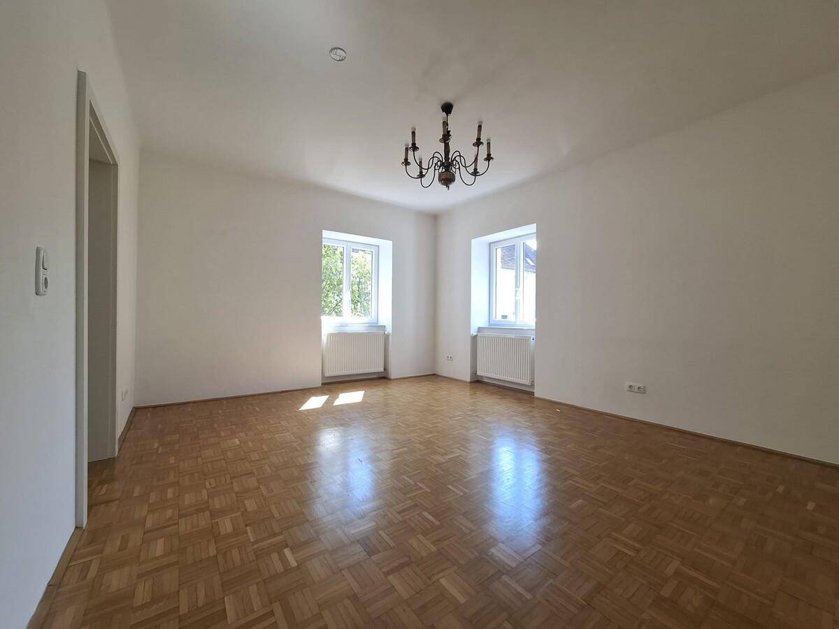 Geräumiges Wohnzimmer mit Parkettboden, hellen Wänden und zwei Fenstern.