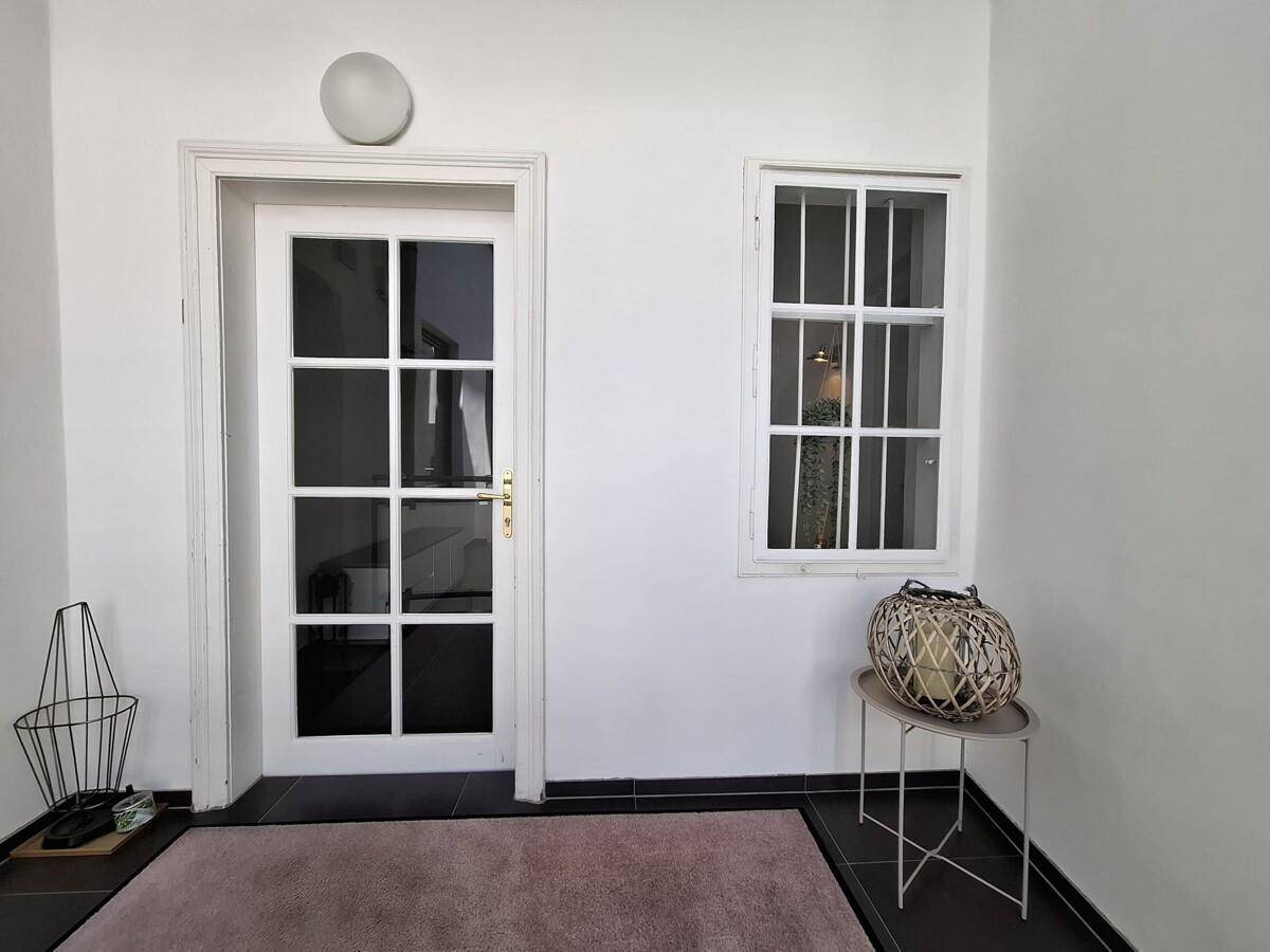 Eingangsbereich mit Glastür, Fenster und dunklen Bodenfliesen, dekoriert mit einem Teppich und Beistelltisch.