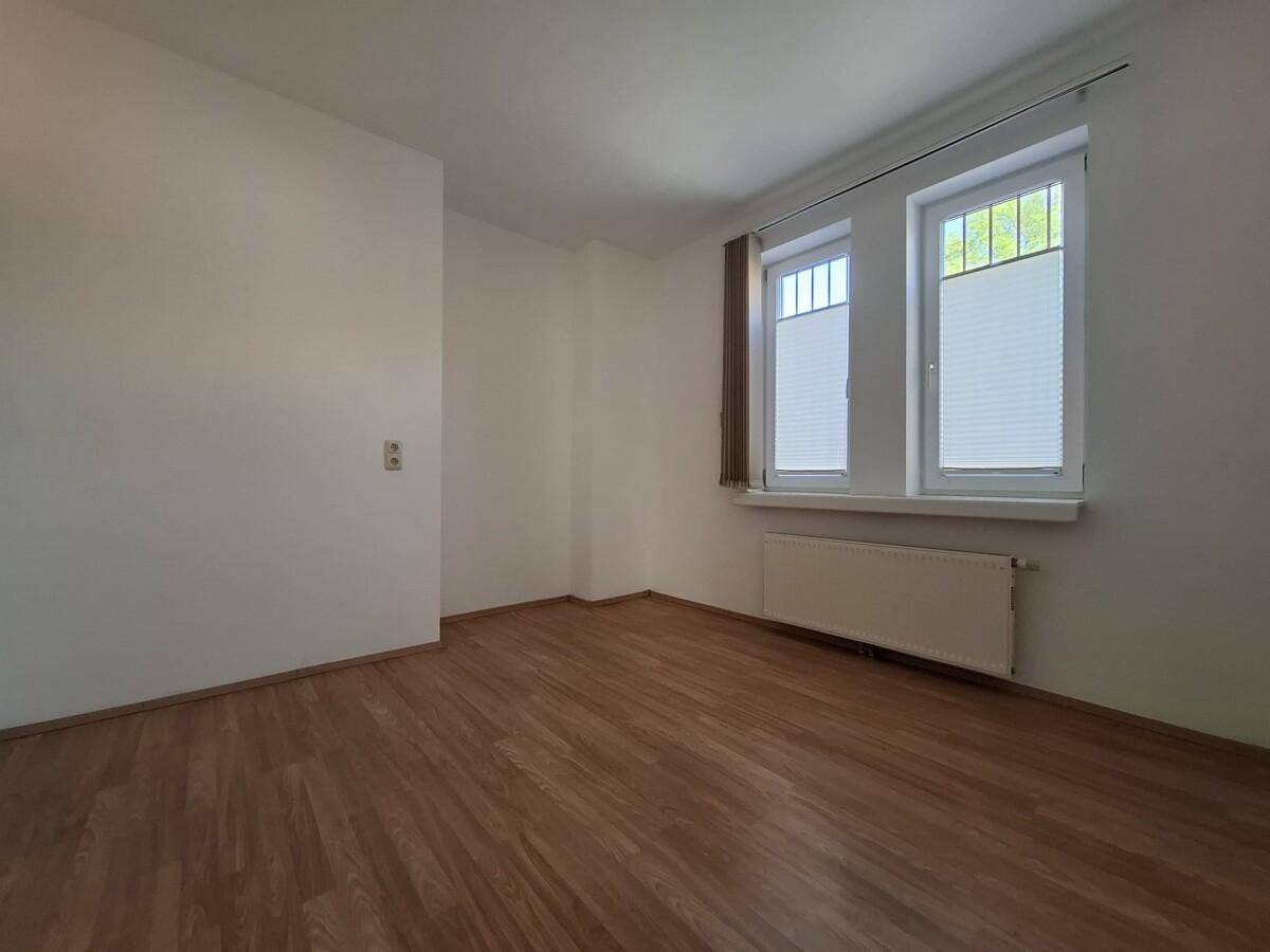 Helles Schlafzimmer mit Laminatboden, zwei Fenstern und Heizkörper unter den Fenstern.