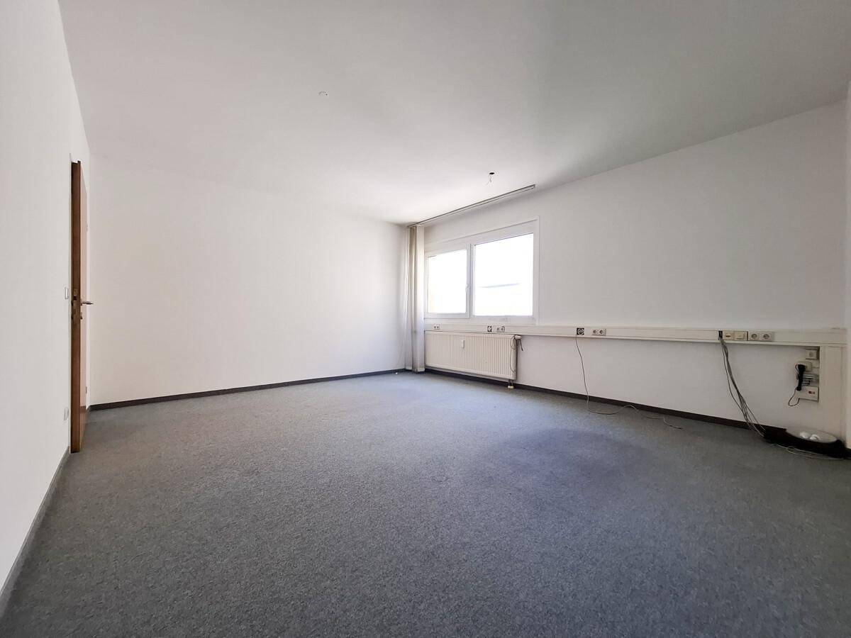 Heller Büroraum mit grauem Teppichboden, einer Holztür und einem Fenster mit Heizkörper.