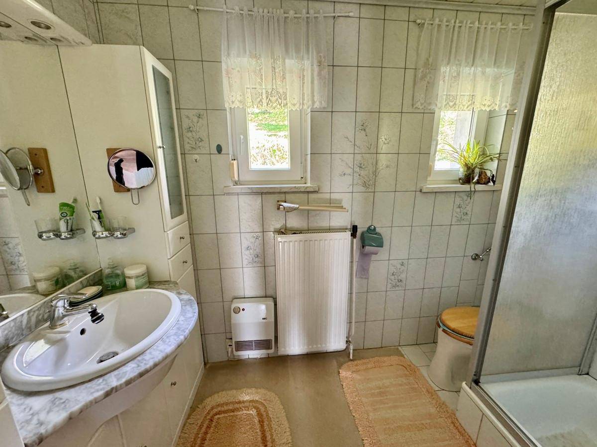 Helles Badezimmer mit Fenster, Dusche, Einzelwaschbecken und Fliesen im 80er-Jahre-Stil.