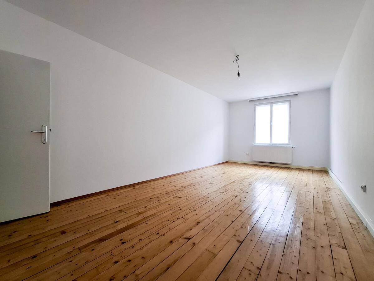 Leeres Zimmer mit Holzdielenboden, weißen Wänden und einem Fenster, das viel Licht hereinlässt.