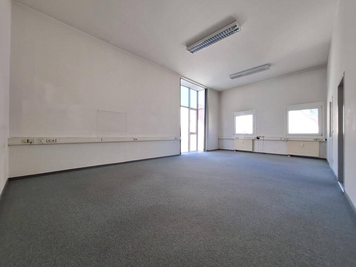Großes, leeres Büro mit Teppichboden und einer hohen Fensterfront, die viel Licht spendet.