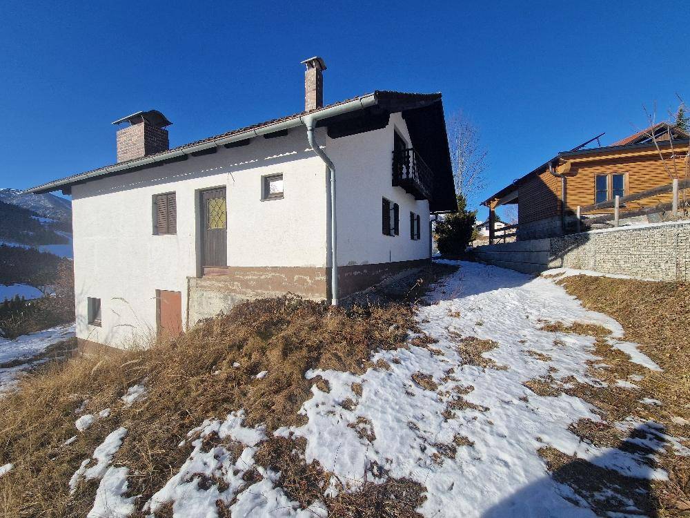 Weißes Haus mit braunem Dach und Fensterläden, umgeben von Schnee und einer Steinmauer.