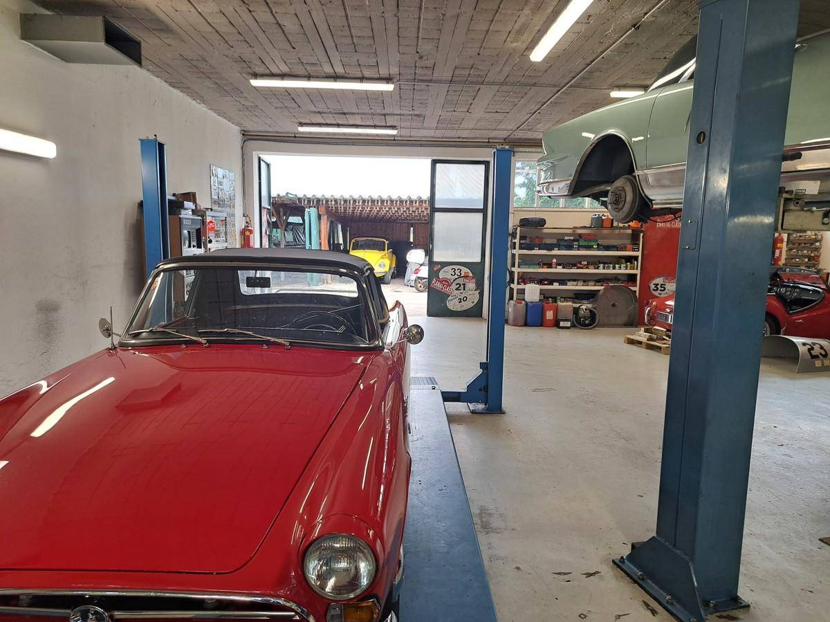 Detailansicht eines roten Oldtimers in der Werkstatt, mit Blick auf den Eingangsbereich und weitere Fahrzeuge.