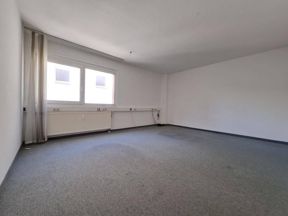 Leerer Büroraum mit grauem Teppichboden, einem Fenster und sichtbaren Kabelkanälen an der Wand.
