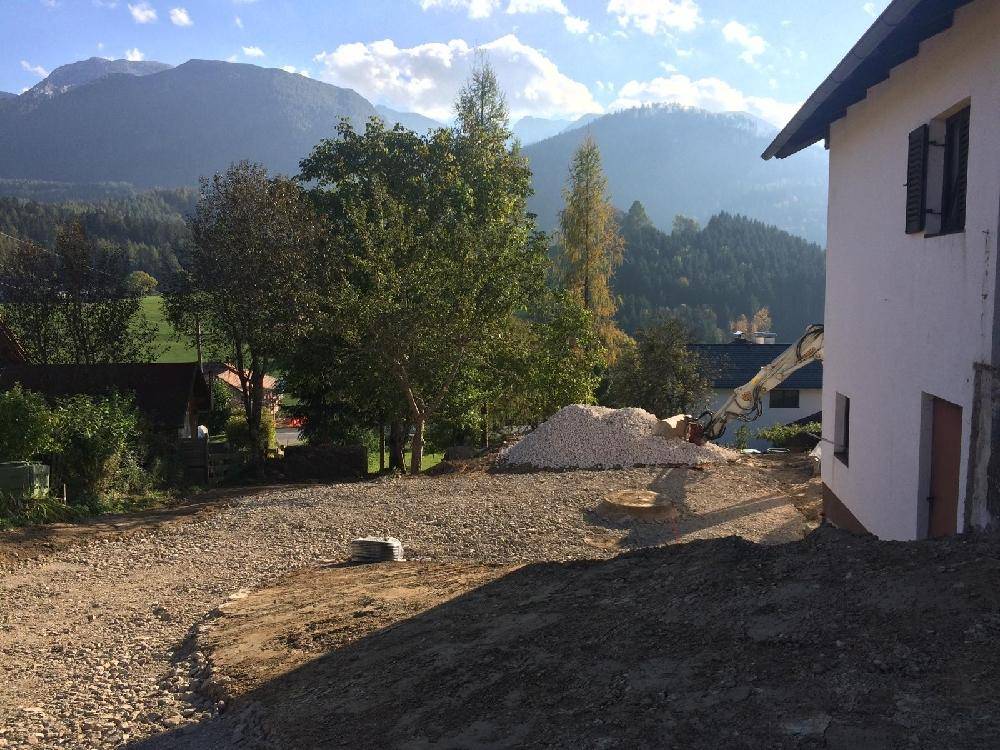 Hausfassade neben einer Baustelle mit Schotterhaufen und Bergen im sonnigen Hintergrund.