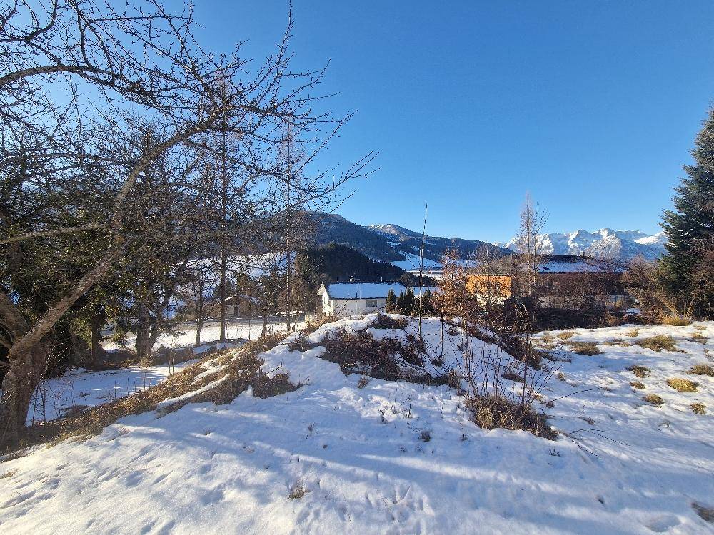 Winterlandschaft mit Schnee, kahlen Bäumen und Häusern vor einer beeindruckenden Bergkulisse unter blauem Himmel.