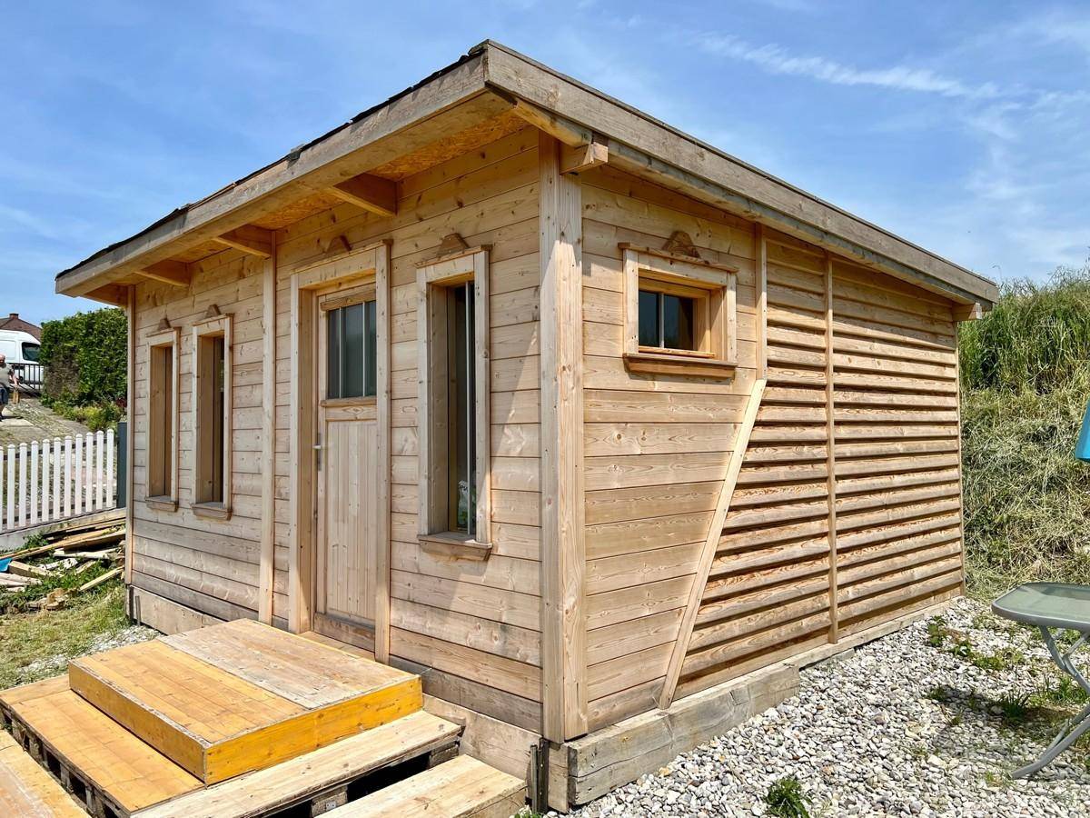 Kleiner Holzschuppen mit Fenstern und einer einfachen Holztür, umgeben von Kies.