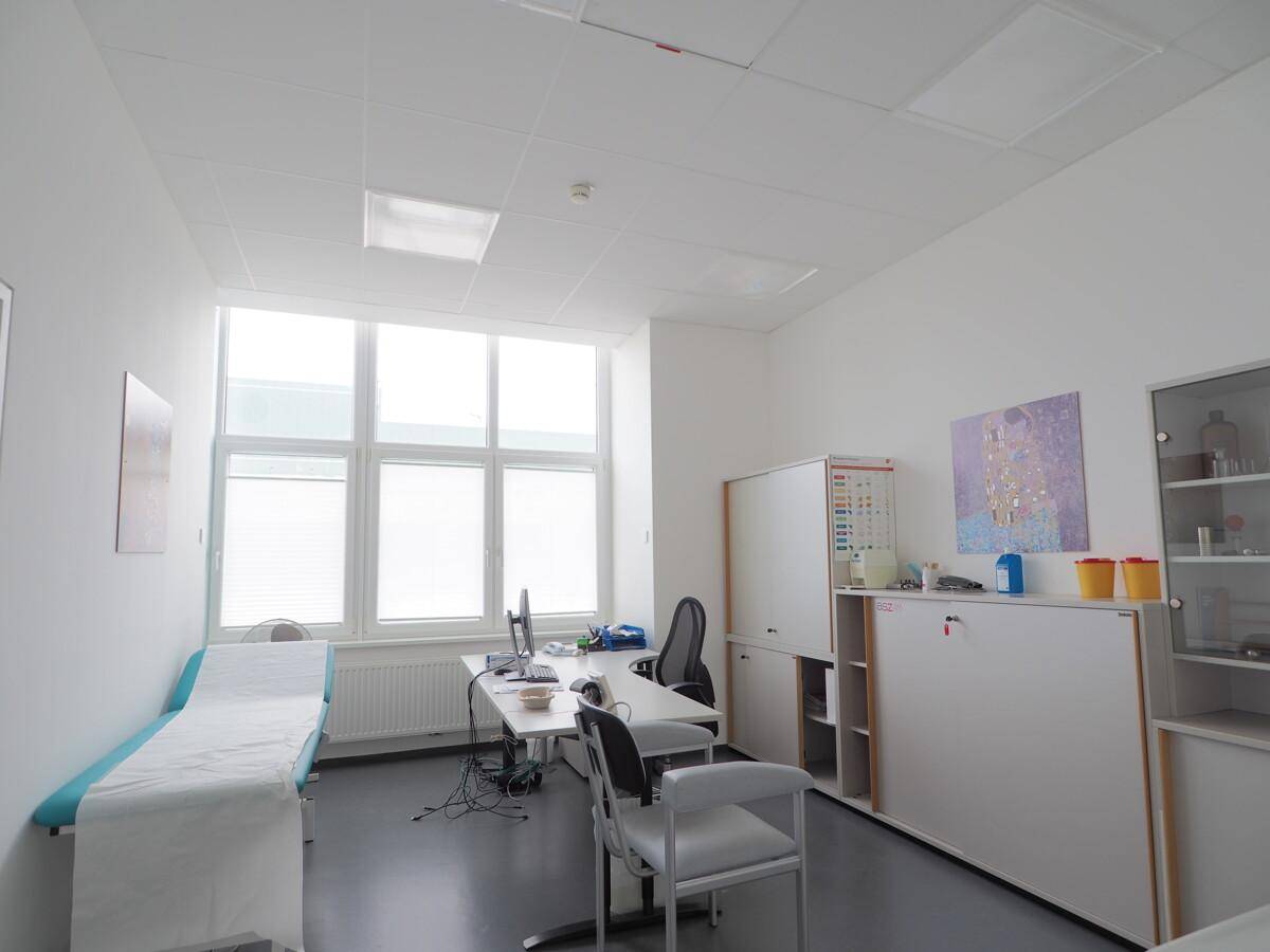 Helles Büro oder Behandlungszimmer mit großem Fenster, Schreibtisch und Untersuchungsliege.