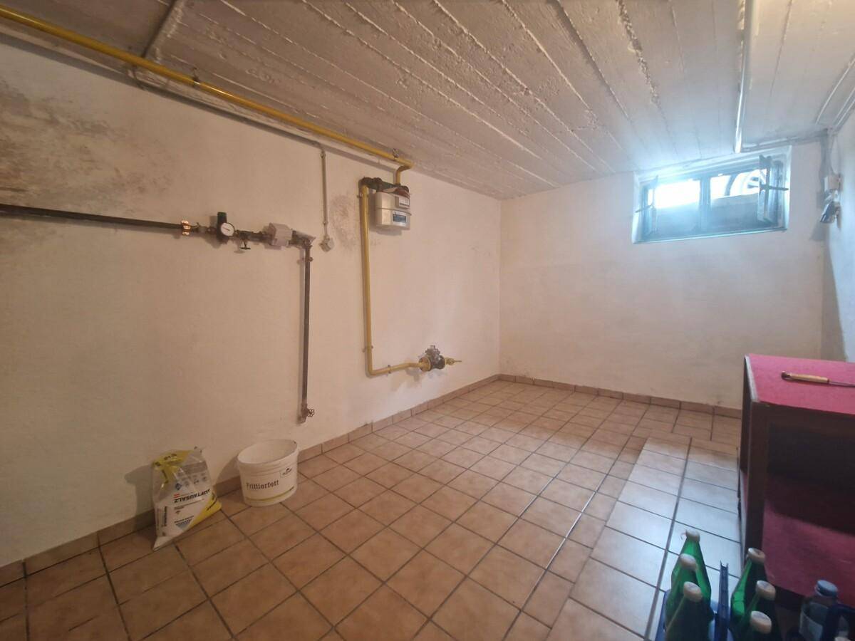 Kellerraum mit Fliesenboden, sichtbaren Leitungen und kleinem Fenster, ideal als Abstellraum.