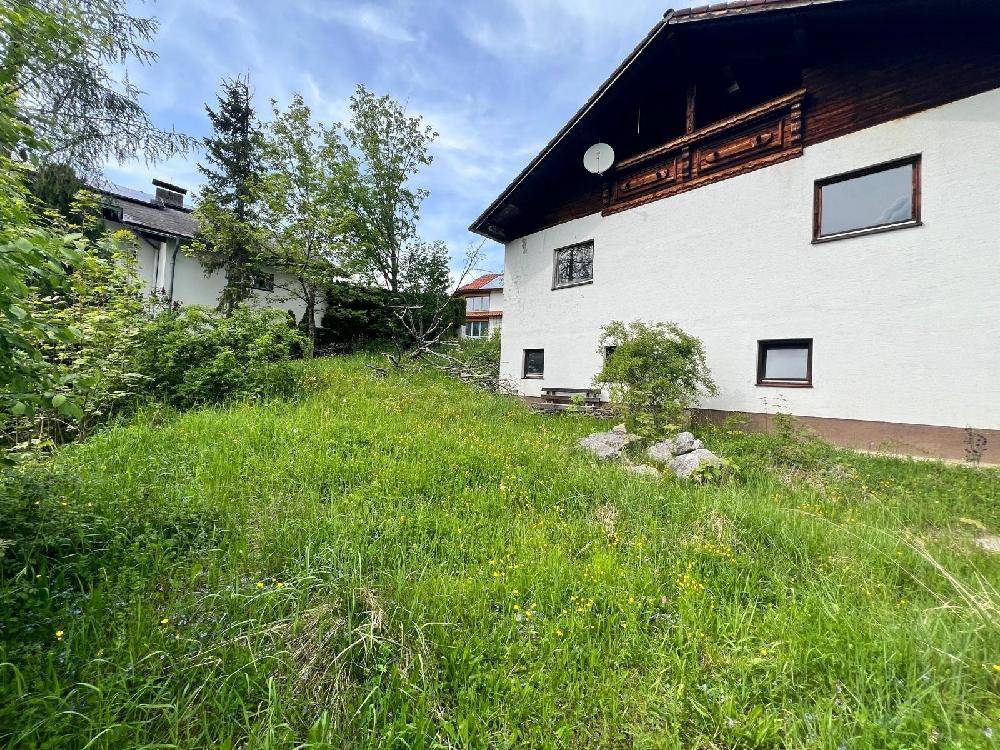 Hausfassade mit weißem Putz und Holzbalkon, umgeben von einer verwilderten Wiese und Bäumen.