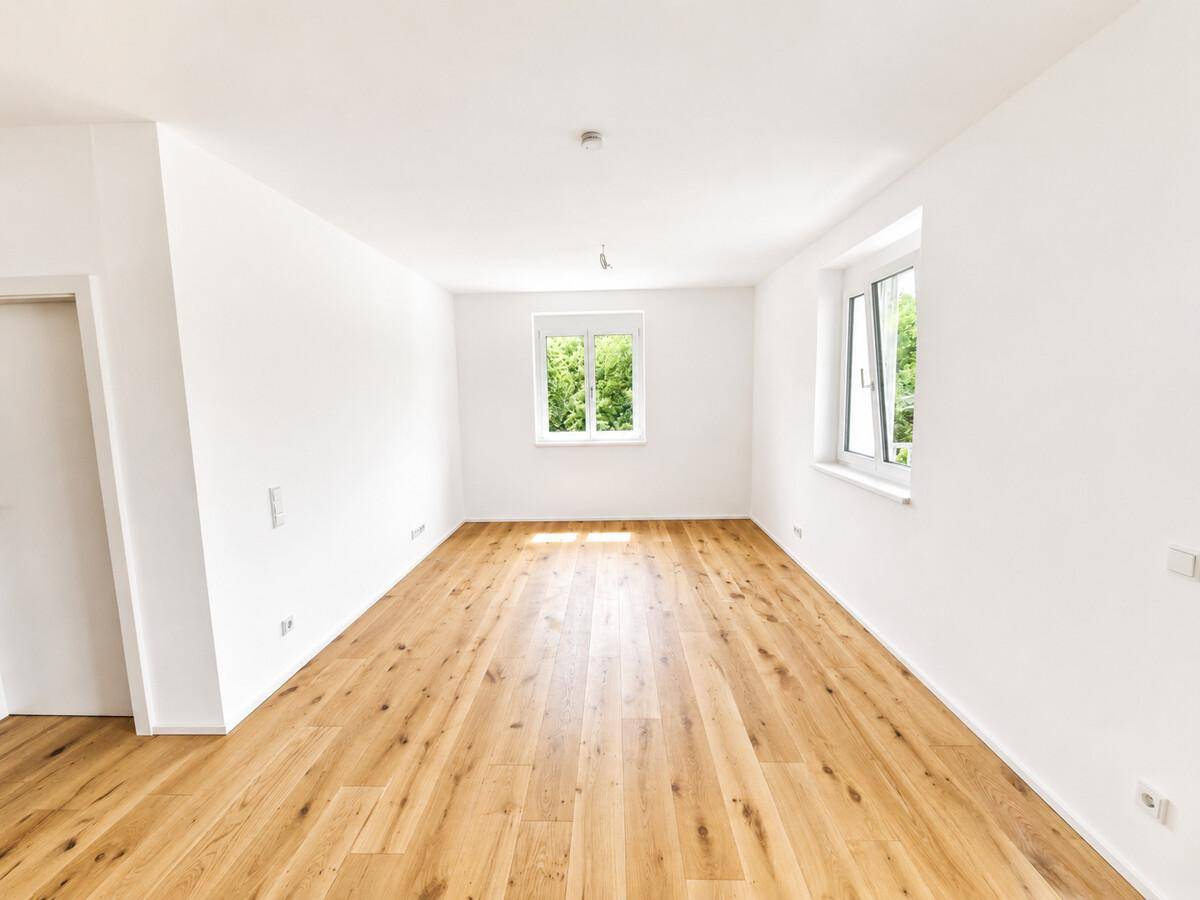 Helles, leeres Wohnzimmer mit neuem Eichenparkettboden und großen Fenstern mit Grünblick.