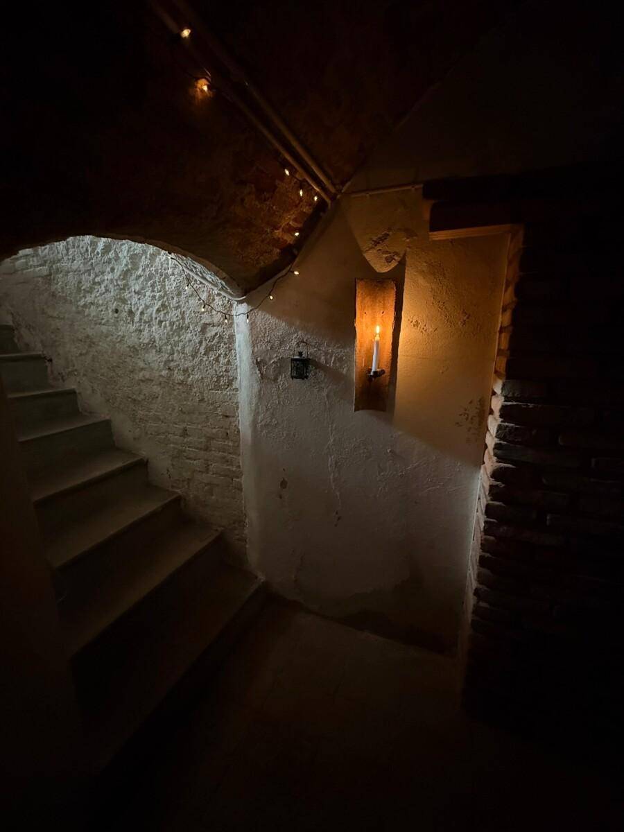 Dunkler Treppenabgang in einen Keller, beleuchtet von einer Kerze und Lichterketten, mit Steinwänden.