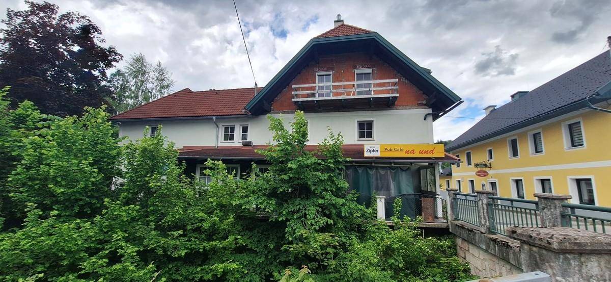 Traditionelles Haus mit Satteldach und Balkon, umgeben von üppigem Grün unter bewölktem Himmel.