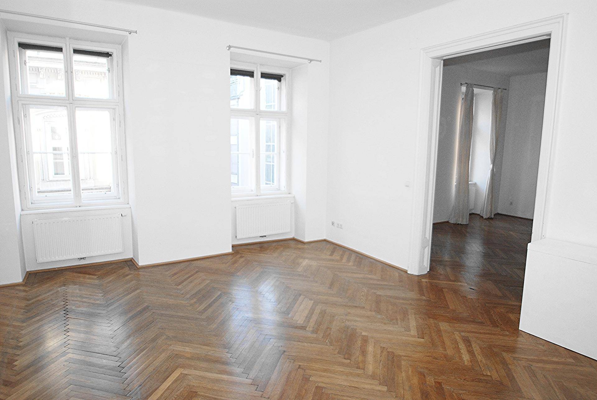 Geräumiges Zimmer mit Fischgrätparkettboden und großen Fenstern, hell und leer.