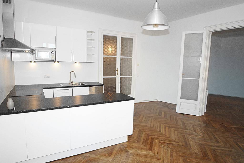 Offene Küche mit weißer Einrichtung und Fischgrätparkett, angrenzend an ein weiteres Zimmer.